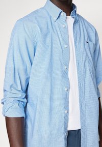 Jasnoniebieska koszula zapinana na guziki z kratkowanym wzorem, długimi rękawami i pojedynczą kieszenią na piersi. Założona na białą koszulkę.