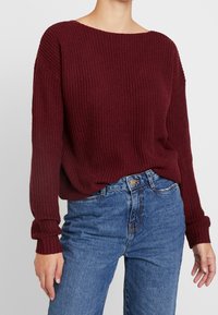 Burgunderfarbener Pullover aus Strick mit weitem Ausschnitt und langen, gerippten Ärmeln. Kombiniert mit hellblauen Jeans aus Denim, die mit einem vorderen Knopfverschluss versehen sind.