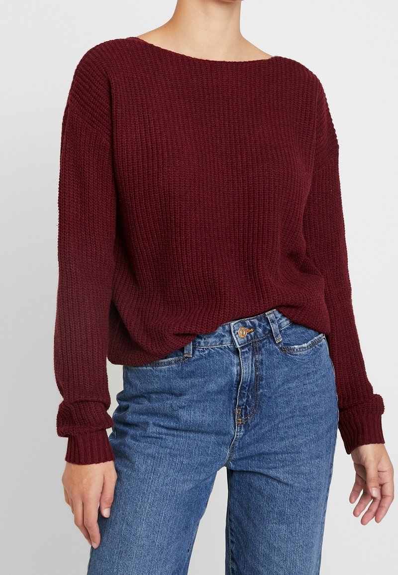 Burgunderfarbener Pullover aus Strick mit weitem Ausschnitt und langen, gerippten Ärmeln. Kombiniert mit hellblauen Jeans aus Denim, die mit einem vorderen Knopfverschluss versehen sind.
