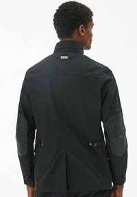 Zwarte jacket met een hoge kraag, textuurstof en leren elleboogpatches. Heeft een rugventilatie en een klein logo detail op de kraag.