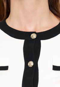 Gros plan sur l'encolure d'un cardigan noir et blanc avec deux boutons décoratifs dorés et une bordure noire autour du col et de la patte de boutonnage.
