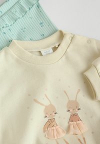 Krémový dětský svetr s ilustracemi dvou králíčků v růžových volánkových sukýnkách. Hladce žebrovaný límec a rukávy, v kombinaci s mátovou texturovanou tkaninou.
