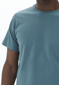 Camiseta de manga corta y cuello redondo en tejido azul apagado. El material parece suave con una textura lisa y tiene costuras mínimas.