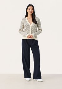 Cardigan rayé în crem și bleumarin, cu decolteu în V, închidere cu nasturi și manșete canelate, asociat cu pantaloni largi bleumarin și pantofi albi.