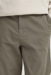 Pantalon en mélange de coton vert olive, avec une texture lisse, des poches latérales et une coupe droite. Les coutures sont minimalement visibles.