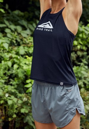 Persoon in zwarte Nike Trail tanktop en grijze Nike shorts die de armen boven het hoofd strekt in een groene, bladerrijke buitenomgeving.