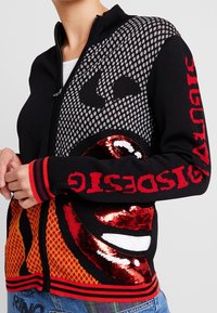 Svart zip-up tröja med stort grafiskt ansiktsdesign, röda paljetterade läppar och röd text på ärmarna, bärs över en vit skjorta.