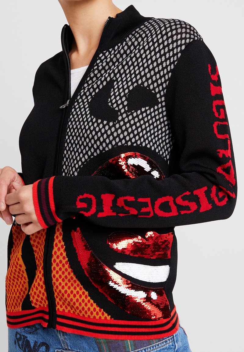 Svart zip-up tröja med stort grafiskt ansiktsdesign, röda paljetterade läppar och röd text på ärmarna, bärs över en vit skjorta.