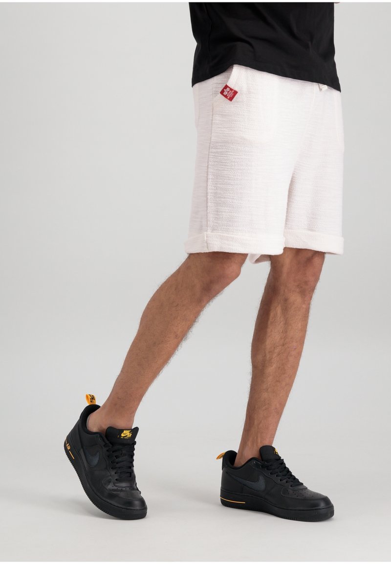 Personne portant un short blanc texturé avec une étiquette rouge et des baskets Nike noires avec des accents jaunes, debout devant un fond uni.