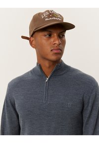 Pulover cu gât zip în lână gri închis, având un guler ribat și un logo brodat subtil. Accesorii cu o șapcă maro pe care este inscripționat un text.