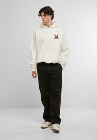 Hanorac cu glugă de culoare crem, cu o buzunar frontal, având un design grafic roșu și negru pe piept. Asortat cu pantaloni închiși la culoare și adidași.