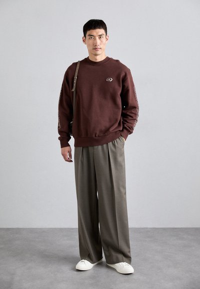 Καφέ φούτερ με λογότυπο στο στήθος, συνδυασμένο με χαλαρές γκρι παντελόνες και λευκά αθλητικά παπούτσια. Το μοντέλο στέκεται με μια τσάντα ώμου.