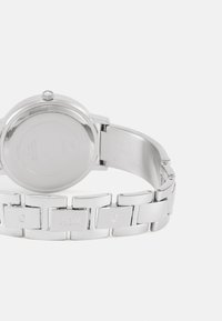 Sølv rustfritt stål klokke med rund urskive, som har en jevn bezel og en lenkebånd preget med "GUESS" og "G" logoer.