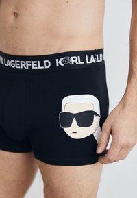 Crne bokserice s bijelim elastičnim pojasom na kojem piše "KARL LAGERFELD." Sadrže grafiku stiliziranog lica s bijelom kosom i sunčanim naočalama.