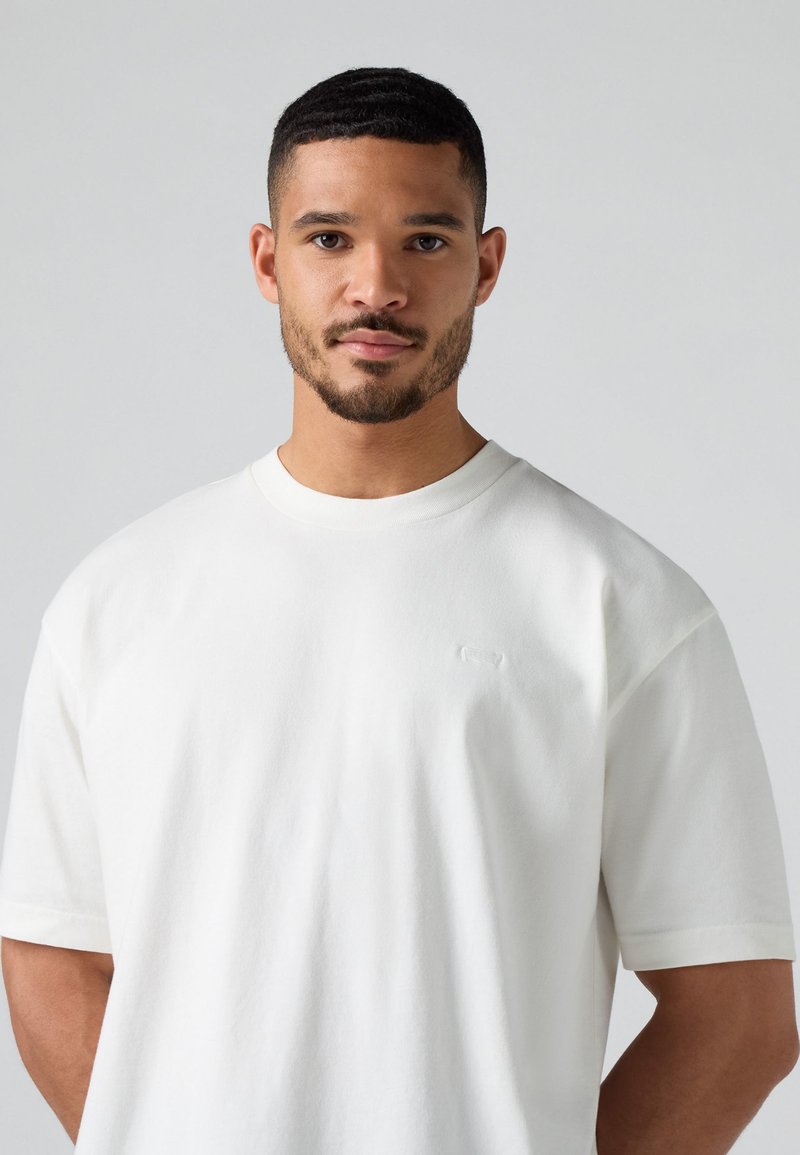 Biała, krótka koszulka z krótkim rękawem, wykonana z miękkiego, gładkiego materiału. Posiada subtelne logo na piersi oraz klasyczny okrągły dekolt.