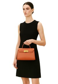 Borsa in pelle arancione con una forma strutturata, manico superiore e chiusura in metallo dorato, tenuta da una persona che indossa un vestito senza maniche nero.