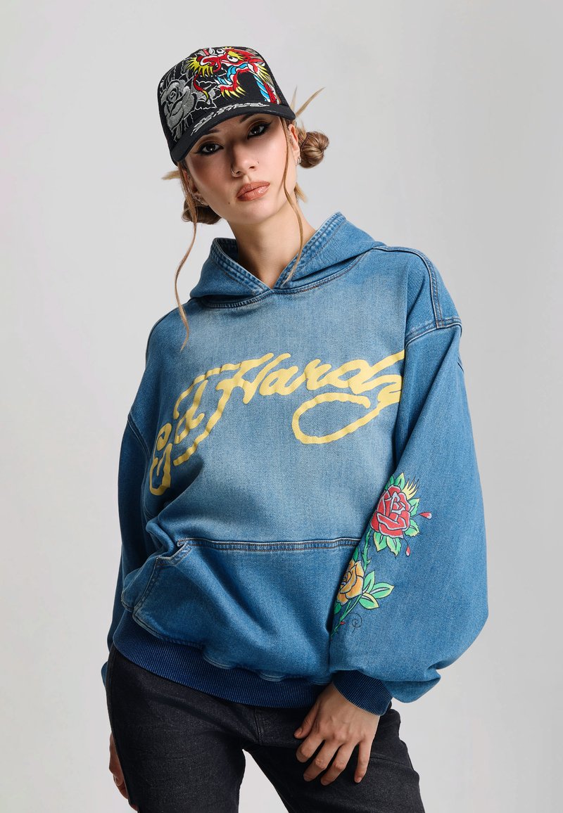 Ung kvinde iført en blå Ed Hardy denim-hoodie med blomsterprint på ærmerne og en sort kasket med et farverigt broderet drage motiv.