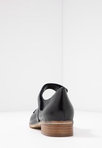 Zapato de cuero negro con corte en la parte trasera, tacón de madera bajo y detalles cosidos a lo largo de los bordes. Superficie suave, forma redondeada.