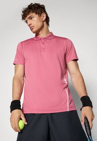 Camisa polo rosa com colarinho clássico, três botões e costura em branco, combinada com calções pretos e pulsos, segurando uma bola de ténis.