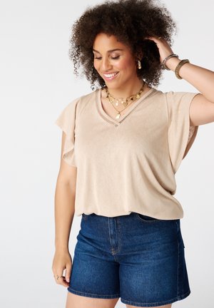 Femme souriante aux cheveux bouclés porte un haut beige à manches courtes, un short en jean bleu et des bijoux en or, regardant vers le bas en touchant ses cheveux avec la main.