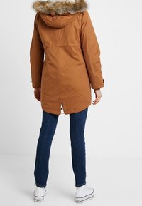 Brown parka jacket with a fur-lined hood, featuring a straight cut, button detailing, and a slit at the back, paired with blue jeans and white shoes.