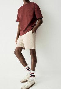 T-shirt bordeaux à manches courtes avec une poche, associé à un short beige. Porte des chaussettes blanches à rayures rouges et bleu marine, ainsi que des baskets crème.