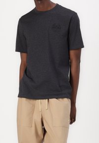 Černé tričko s krátkým rukávem z měkké látky, s nenápadným kruhovým logem na hrudi. Nošeno s béžovými kalhotami.