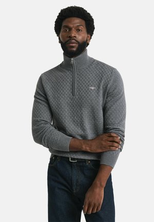 Sudadera gris con cremallera, con un patrón de cuadros texturizado y puños acanalados. Combinada con jeans azul oscuro.