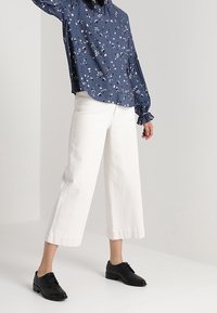 Marinblus med blommönster, långa ärmar och samlade manschetter, parat med vita vida byxor. Svarta snörskor fulländar outfiten.