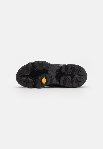 Suela de goma negra con surcos profundos y un destacado logo de Vibram en amarillo. Superficie texturizada para tracción y durabilidad.
