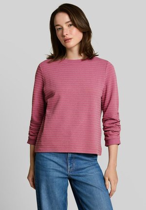 Femme aux cheveux bruns mi-longs portant un t-shirt rose à manches longues rayé et un jean bleu, debout devant un fond clair uni.