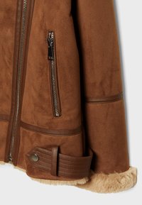 Ein brauner Wildlederjackenärmel mit Reißverschlusstasche, Lederbesatz, Riemen mit Druckknopf und sichtbarem beigem Schaffellfutter am Bündchen und Saum.