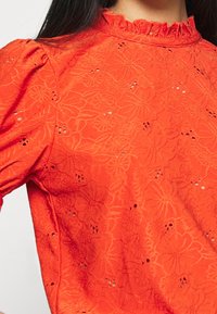Blusa rossa ricamata con motivo floreale, dotata di maniche a sbuffo e collo arricciato. Il tessuto presenta una finitura testurizzata e piccoli dettagli a intaglio.