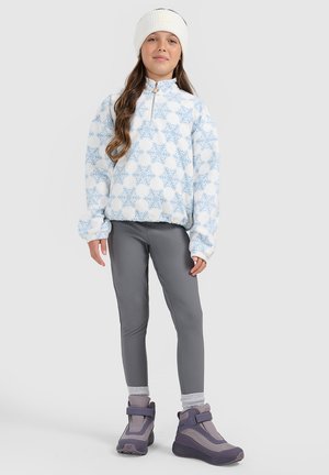 Jong meisje met een witte muts, blauwe fleece trui met sneeuwvlokpatroon, grijze legging, grijze sokken en paarse winterlaarzen, staand op een witte achtergrond.