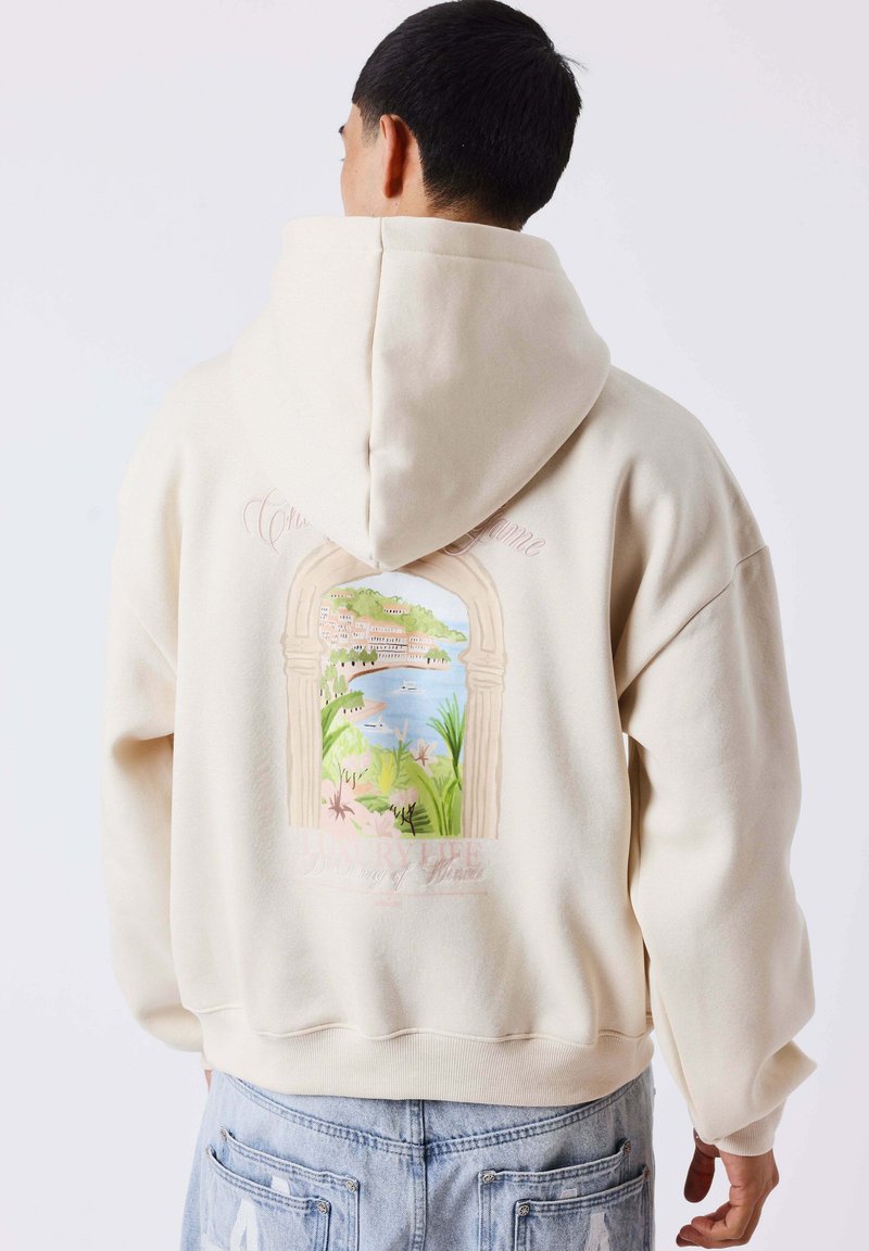 Homme portant un sweat à capuche crème avec une illustration colorée en forme d'arche d'une ville côtière et de verdure dans le dos.