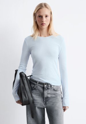 Young woman with blonde hair wearing a light blue long-sleeve top and gray jeans, holding a black leather bag by her side.