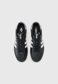 Black leather sneakers with three white stripes, a textured green rubber sole, and a silver zip detail on the upper.