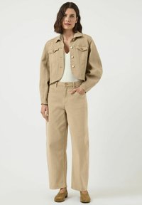 Vrouw met schouderlang bruin haar, gekleed in een beige cropped jack, hooggewaisted beige broek, witte top en bruine schoenen, staande tegen een effen achtergrond.