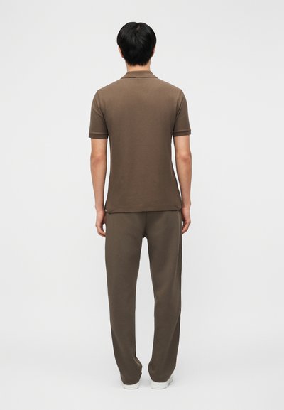 Άνδρας στέκεται πλάτη φορώντας καφέ κοντομάνικο πόλο πουκάμισο και καφέ παντελόνια, με λευκά παπούτσια, σε απλό φόντο.