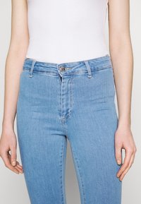 Jeans azul claro com um design ajustado, apresentando um fecho com zip e botão, passadores de cinto e discretos detalhes de costura ao longo das costuras.