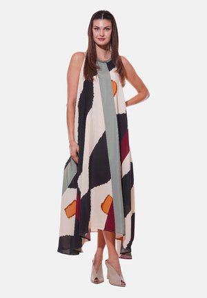 Donna con lunghi capelli castani che indossa un vestito lungo senza maniche, fluente, con motivi astratti neri, bianchi, grigi, arancioni e bordeaux, e tacchi beige.