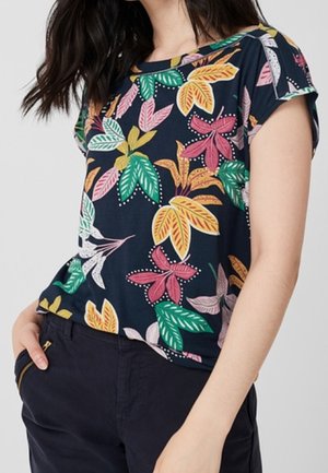 Kobieta ubrana w granatową kwiecistą koszulę z krótkim rękawem z zielonymi, żółtymi i różowymi liśćmi, wpuszczoną w ciemnogranatowe spodnie z kieszeniami na zamek.