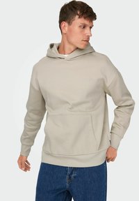Felpa con cappuccio beige realizzata in tessuto morbido, con tasca frontale, polsini a coste e un logo discreto sul petto. Indossata con jeans in denim.