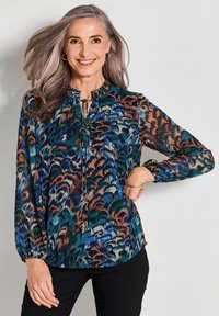 Blusa multicolore in tessuto leggero con un vivace motivo astratto verde, blu, arancione e marrone. Collo arricciato con lacci.