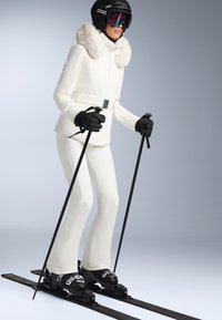 Fehér síruha beépített, szőrmebéléses kapucnival, fekete kesztyűk és síléc. Fekete sícipők ezüstszínű fémrészekkel, letisztult dizájnnal és sima textúrával.