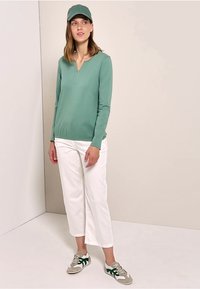 Helles grünes Langarmoberteil mit V-Ausschnitt, kombiniert mit weißen verkürzten Hosen und grau-grünen Sneakern. Das Outfit umfasst auch eine passende Mütze.