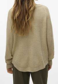 Maglione beige lavorato a maglia con motivo strutturato, spalle scese e orlo arrotondato, abbinato a pantaloni verde oliva, visto da dietro.