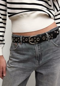 Zwarte leren riem met zilverkleurige beslag, voorzien van een reeks ronde oogjes en een metalen gesp, gedragen over grijze denim jeans.