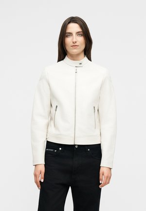 Γυναίκα με ίσια καστανά μαλλιά φοράει εφαρμοστή λευκή ζακέτα με φερμουάρ και ψηλό γιακά, μαζί με μαύρο παντελόνι, στέκεται μπροστά σε μονόχρωμο φόντο.