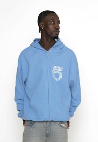 Felpa con zip di colore azzurro chiaro, tessuto testurizzato, tasche anteriori e testo grafico bianco sull'area sinistra del petto. Presenta un cappuccio.
